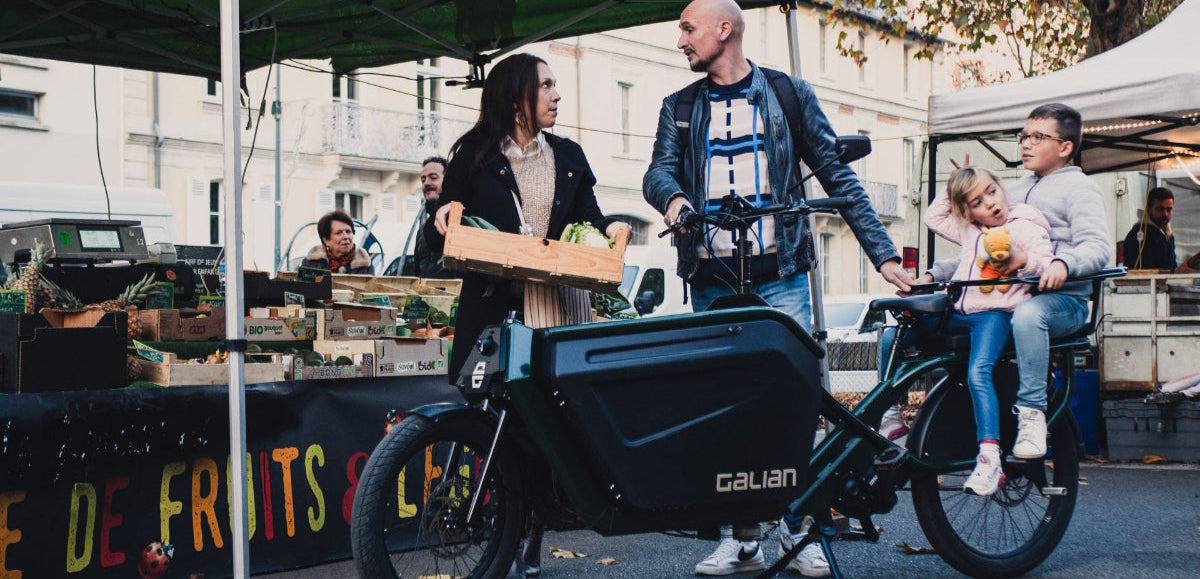 deux adultes et deux enfants dans un velo cargo au marche